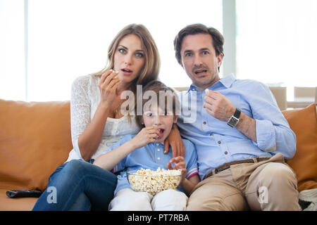 Bella giovani genitori e il loro figlio stanno guardando la TV, mangiare popcorn seduti sul divano di casa Foto Stock
