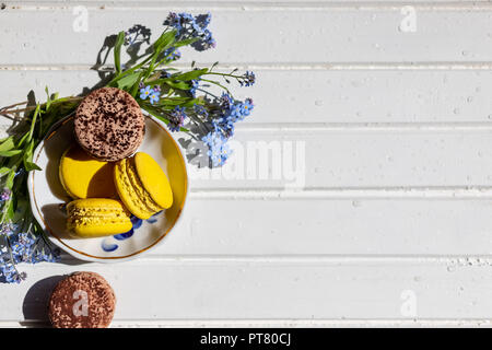 Giallo delizioso cioccolato e amaretti sul trendy bianco sullo sfondo di legno con fiori di primavera, piatto laici. gustoso giallo e marrone amaretti. spazio per il testo. candy per la festa il dessert e dimenticare me not e narcisi Foto Stock