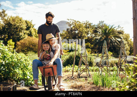 Padre dà la madre e la figlia ride in carriola presso l'azienda. La famiglia felice con l uomo che donna e bambina ride in carriola all'esterno. Foto Stock