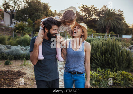 Allegro giovane famiglia di trascorrere del tempo insieme alla fattoria. I giovani genitori con la loro piccola figlia all'aperto nel campo. Poco ragazza seduta su fathe Foto Stock