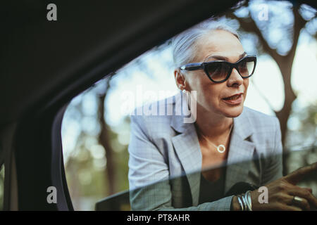 Bella donna senior parlando con taxi driver attraverso apre la finestra auto. Imprenditrice in occhiali da sole a parlare con cabina conducente. Foto Stock