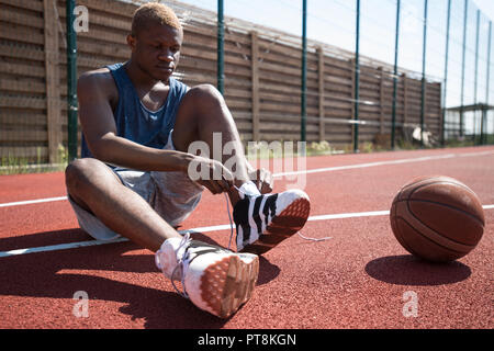 A piena lunghezza ritratto della moderna africana del giocatore di basket scarpa legatura seduto sul pavimento nella corte esterna, spazio di copia Foto Stock