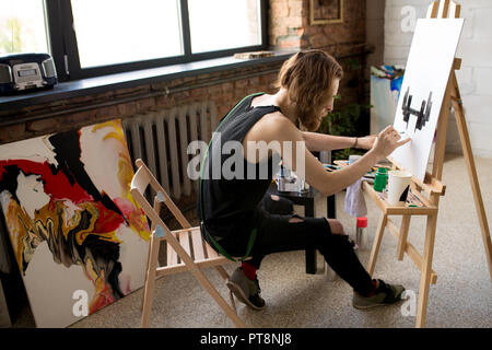 Angolo alto ritratto di capelli lunghi artista contemporanea pittura immagine sul cavalletto in inchiostro nero mentre godendo il lavoro in art studio, spazio di copia Foto Stock