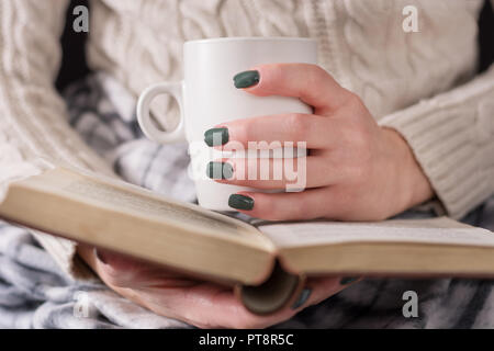 Ragazza detiene il libro bianco e la tazza di tè in mani. Donna indossa un maglione di lana e coperto con una coperta. Il freddo inverno e concetto. Close up, il fuoco selettivo Foto Stock