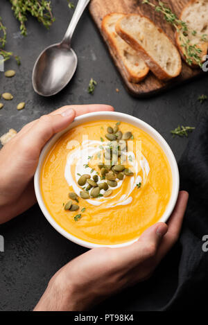Persona in possesso di una ciotola di zuppa di zucca nero su sfondo concreto, vista dall'alto Foto Stock