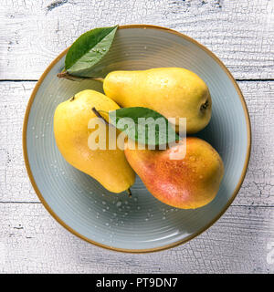 Immagine quadrata di wet mature di colore giallo dorato pere e foglie verdi sul round piastra blu su bianco antico tavolo in legno superficie. Messa a fuoco selettiva. Vista da sopra. Foto Stock