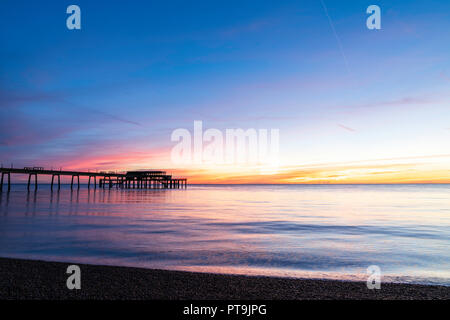 Trattare pier con un mare calmo e il sorgere del sole sul mare orizzonte in un cielo parzialmente chiara e in parte con soffice nuvola di sottili strati. Foto Stock