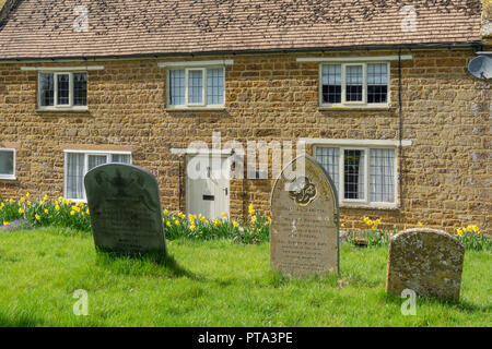 Vista dal sagrato della chiesa di Sant'Andrea, lapidi in primo piano, vecchio cottage in pietra a posteriori; il villaggio di vecchi, Northamptonshire, Regno Unito Foto Stock