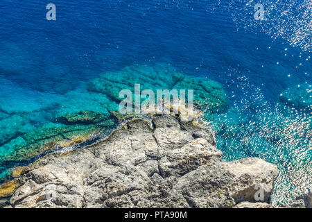 Foto orizzontale con la vista verso il basso sulla scogliera rocciosa con il mare blu intorno a. Il reef è sotto l'acropoli di Rodi città Lindos catturato nella soleggiata giornata estiva. Foto Stock