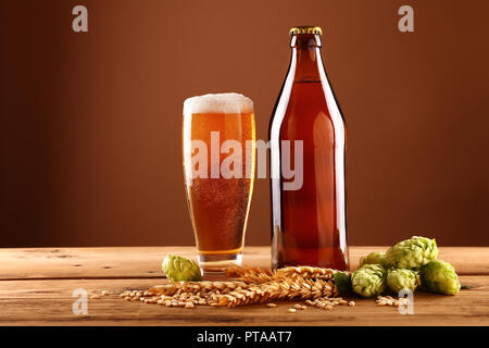 Chiudere completamente una bottiglia marrone e un bicchiere di birra con la schiuma e bolle, verde luppolo, orzo e grano spike sul tavolo di legno, basso angolo vista laterale Foto Stock