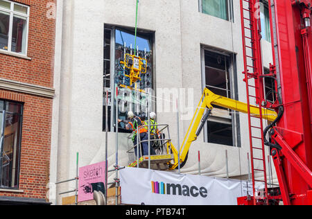 Impresa edile macis lavoratori su un cherry picker paranco con attenzione il montaggio di un nuovo grande finestra in vetro lastra in un edificio a Mayfair, London, Regno Unito Foto Stock