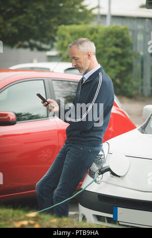 Uomo che utilizza il telefono cellulare mentre si ricarica auto elettrica Foto Stock