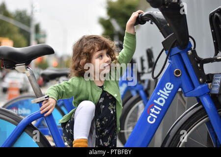 Un impavido piccola ragazza con i capelli ricci si arrampica su una Citibike con uno sguardo di determinazione nel suo occhio, pur non sapendo come andare in bicicletta. Foto Stock