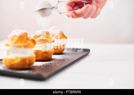 Spolverata di zucchero a velo su una deliziosa crema torte di sfoglia su un piatto nero su bianco tavola di marmo su sfondo rosa. Messa a fuoco selettiva, copia dello spazio. Foto Stock
