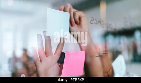 Mani di donna che incollano appunti appiccicosi su una parete di vetro in ufficio. uomini d'affari che discutono nuove idee pubblicando appunti it su una parete di vetro trasparente. Foto Stock