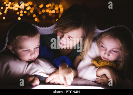 Madre e bambini al di sotto della coperta utilizzando digitale compressa contro le luci di Natale Foto Stock