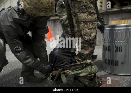 Una massa giapponese di autodifesa membro di servizio consente di U.S. Lancia Marine Cpl. Enrique Puentes Jr. uscire dalla sua missione orientata protettivi tuta di postura durante una simulazione di studio congiunto al Marine Corps Air Station Futenma, Okinawa, in Giappone, 13 luglio 2017. Il servizio JGSDF membro è con xv nucleare, biologico, chimico unità di difesa. Puentes, nativo di Tampa, Florida, è una difesa CBRN specialista con ala Marine Headquarters Squadron 1. Foto Stock