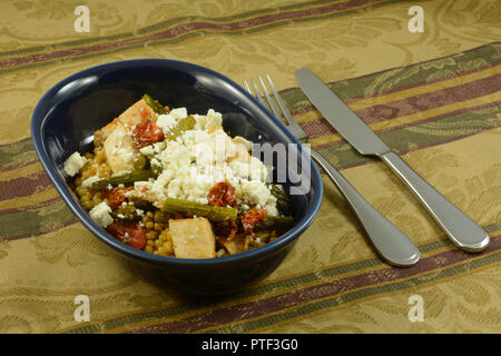 Il cous cous israeliano o ptitim coperti con pollo e verdure arrosto di asparagi e pomodori con il formaggio feta sulla parte superiore di forma ovale piatto blu Foto Stock