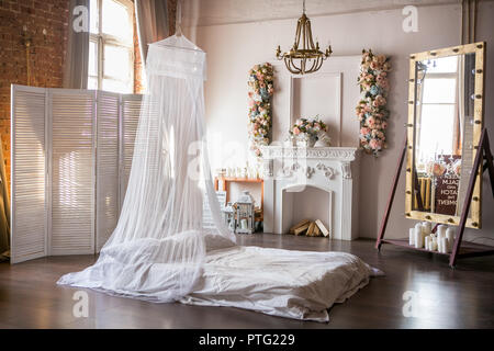 Stile loft a camera con un letto a baldacchino, un caminetto bianco con una disposizione del fiore, uno schermo bianco, un grande specchio e le candele nel pomeriggio Foto Stock