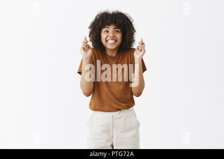 Tiro al coperto di affascinanti sognanti ottimista di carnagione scura femmina marrone in t-shirt scrollare dall impazienza sorridente largamente incrocio le dita per la buona fortuna, in attesa di sogno oltre il muro grigio Foto Stock