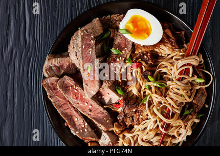 Close-up di nero ciotola di Soba Noodles con fette di arrosto di manzo, funghi shiitake, metà di uovo sodo e verdure fritte con bacchette, asiatica Foto Stock