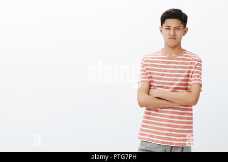 Carino giovane ragazzo cinese in striped t-shirt cercando offeso e mad, mantenendo un insulto all'interno, il broncio attraversando le mani contro il petto e accigliata in piedi sul lato destro dello spazio di copia Foto Stock