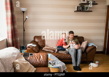 Il padre e i suoi due figli seduti sul divano del soggiorno a casa spendendo un po' di tempo insieme. Il ragazzino è sempre sorridente e guardare la tv whil Foto Stock