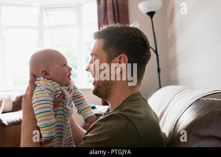 Padre tenendo la sua new born baby boy fino al suo volto a fare di lui ridere. Sono seduto in soggiorno sul divano. Foto Stock