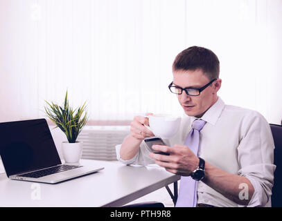 Giovane imprenditore commerciante si siede a un tavolo al mattino in una stanza vicino a un computer portatile con una tazza di caffè. Foto Stock