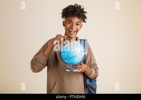 Ritratto di un giovane positivo afro american uomo vestito di felpa con cappuccio zaino porta isolata, puntando al globo terrestre Foto Stock