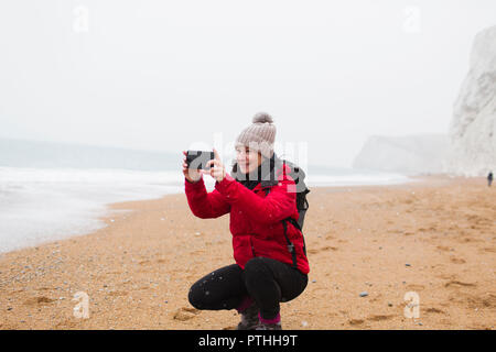 Donna in abiti caldi tramite telefono cellulare dotato di fotocamera digitale sulla spiaggia innevata Foto Stock