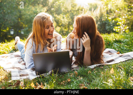 Immagine di due giovani donne che indossano abiti casual giacente su erba nel parco e utilizzando argento laptop Foto Stock