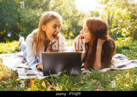 Immagine di due entusiasti le donne indossano abiti casual giacente su erba nel parco e utilizzando argento laptop Foto Stock