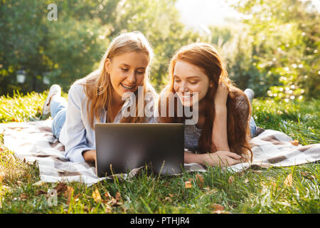 Immagine di due bella femmina amici indossando abiti casual giacente su erba nel parco e utilizzando argento laptop Foto Stock