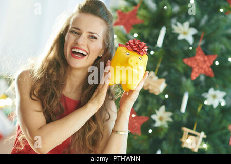 Felice alla moda donna in abito rosso giallo di scuotimento salvadanaio con fiocco rosso nei pressi di albero di Natale Foto Stock