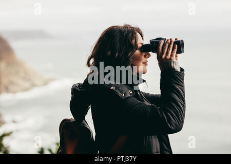 Vista laterale di un escursionista femmina cercando in binocolo godendo di una vista spettacolare dalla cima della montagna. Donna che indossa una giacca calda guardando attraverso il binocolo. Foto Stock