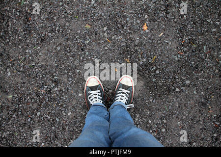 Guardando giù su un paio di gambe in jeans che indossano converse in piedi sul marciapiede. Foto Stock