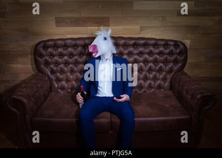 Funny unicorn in elegante abito si siede sul divano con una bottiglia di vino. Uomo insolito con il mal di testa a casa. Freaky boss in maschera comica. Foto Stock