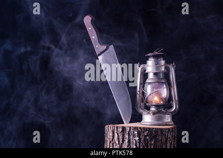 Coltello di grosse dimensioni e la combustione di olio vecchio lampada sul registro di legno nella foresta nera e sfondo velato. L' orrore e la nozione di Halloween Foto Stock