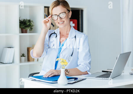 Sorridente femmina adulta del medico la regolazione degli occhiali a tavola con il computer portatile in ufficio Foto Stock