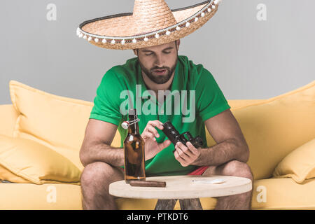 Uomo delinquente in sombrero messicano sul divano giallo con la pistola, un sigaro e una bottiglia di birra su grigio Foto Stock