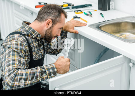 Barbuto repairman tenendo una chiave regolabile e guardando sotto il lavandino rotti in cucina Foto Stock