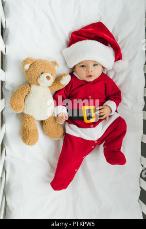 Vista superiore del bellissimo piccolo bambino in santa suit giacente nella mangiatoia con Teddy bear Foto Stock
