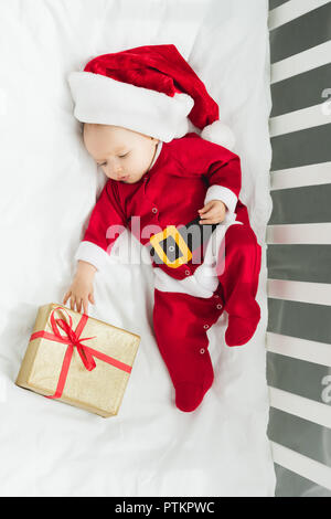 Vista superiore del bellissimo piccolo bambino in santa suit giacente nella mangiatoia con un regalo di natale Foto Stock