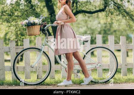 Ritagliato colpo di donna elegante in abito con bicicletta retrò con cesto in vimini pieno di fiori in campagna Foto Stock