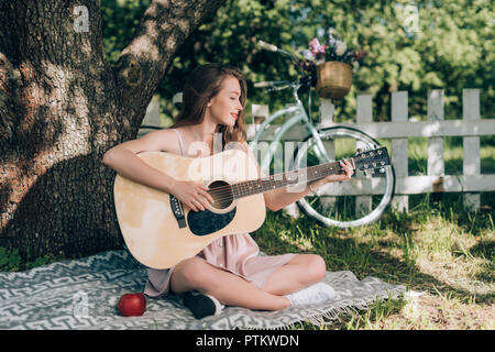 Attraente giovane donna suonare la chitarra acustica mentre poggia su una coperta sotto agli alberi in campagna Foto Stock