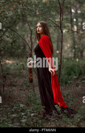 Attraente donna mistica in abito nero e mantello rosso azienda violino in foresta Foto Stock