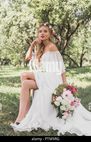 Giovane e bella sposa in ghirlanda floreale e abito da sposa seduti su una sedia e guardando la fotocamera all'aperto Foto Stock