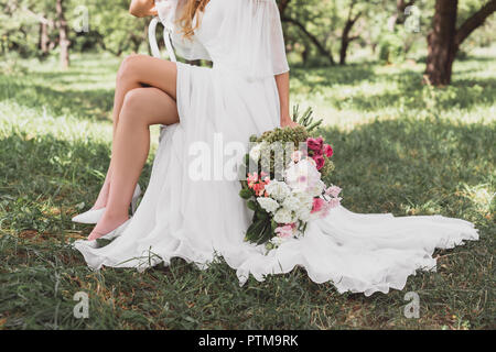 Ritagliato colpo di sposa in abito da sposa seduti su una sedia e tenendo premuto bouquet di fiori all'aperto Foto Stock