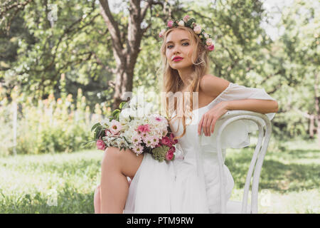 Bella bionda e giovane sposa con abito da sposa seduti su una sedia e guardando la fotocamera all'aperto Foto Stock